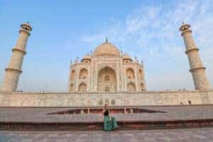 A girl sitting in front of the Taj Mahal, surrounded by a few people, under a clear blue sky. The iconic white marble monument is visible in the sunlight, showcasing its intricate architectural details.