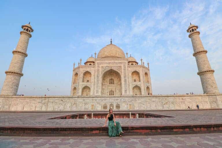 A girl sitting in front of the Taj Mahal, surrounded by a few people, under a clear blue sky. The iconic white marble monument is visible in the sunlight, showcasing its intricate architectural details.