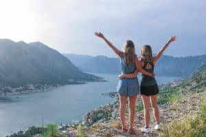 Two solo travellers who became friends through conversation started by travel questions. overlooking Kotor Bay. There is a large blue water body surrounded by a mountainous landscape. The sky is blue with some clouds.