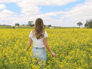 A young woman with long brown hair, wearing a white shirt and light blue jeans, standing in a vibrant yellow canola field under a partly cloudy sky.