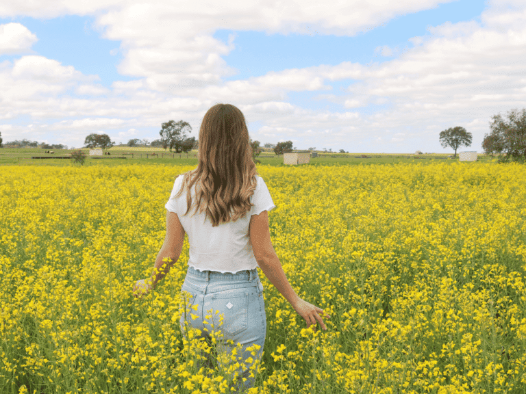 A young woman with long brown hair, wearing a white shirt and light blue jeans, standing in a vibrant yellow canola field under a partly cloudy sky.