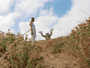 A woman wearing light-colored clothing stands on a dirt path surrounded by dry grass and wildflowers, gazing at a whimsical sculpture of a creature with raised hands, set against a bright blue sky with fluffy clouds.
