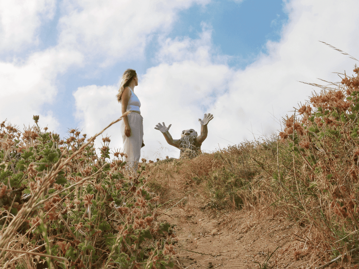 A woman wearing light-colored clothing stands on a dirt path surrounded by dry grass and wildflowers, gazing at a whimsical sculpture of a creature with raised hands, set against a bright blue sky with fluffy clouds.