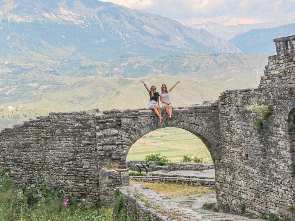 Two travellers sitting on an ancient stone archway in a historic castle ruin, surrounded by lush green hills and mountains in Albania. They are smiling and raising their arms in a celebratory pose.