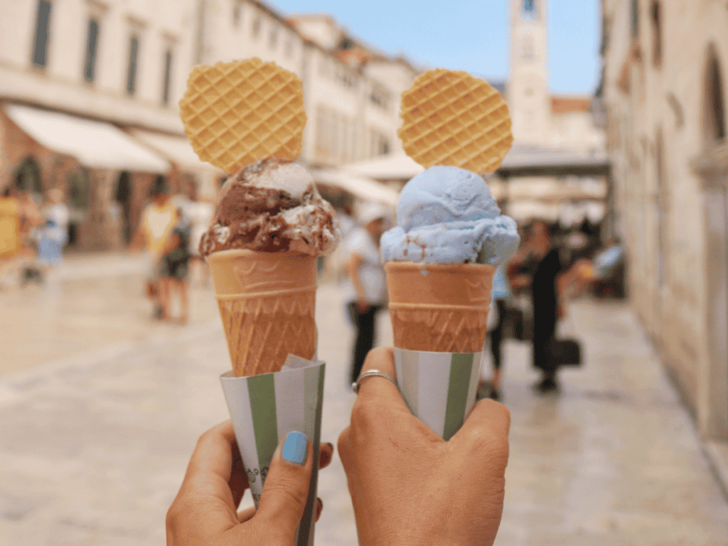 Two ice cream cones being held up in a cobblestone street of Dubrovnik, Croatia. The ice cream flavours include Neapolitan and bubblegum, with waffle toppings, and the background shows historic European architecture and a clock tower.